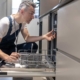 A technician kneels to inspect an open kitchen dishwasher while troubleshooting potential underlying plumbing and drainage problems for repair.
