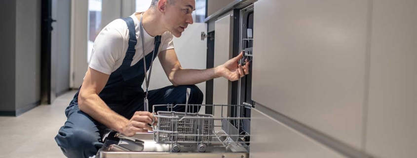 A technician kneels to inspect an open kitchen dishwasher while troubleshooting potential underlying plumbing and drainage problems for repair.