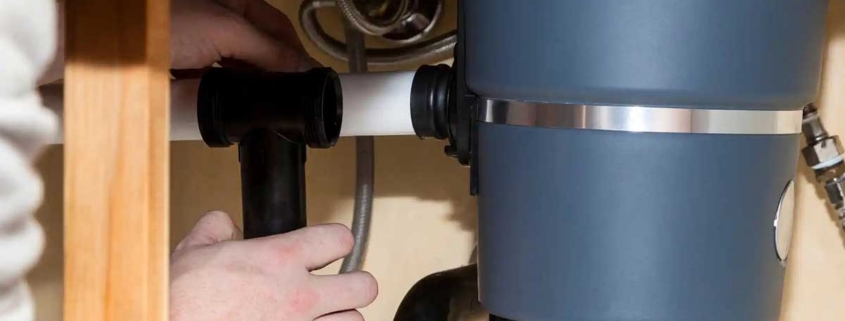 Hands work under a kitchen sink to inspect and repair plastic pipe connections causing a garbage disposal unit to leak.
